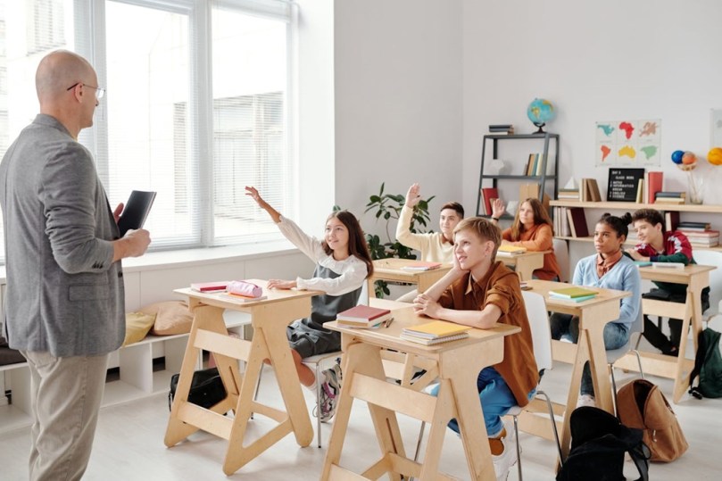 Teacher at front of classroom with children seated at desks, some raising their hands