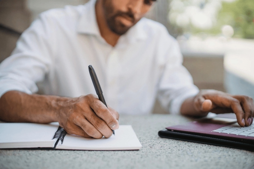 A person writing on a notebook while using his laptop