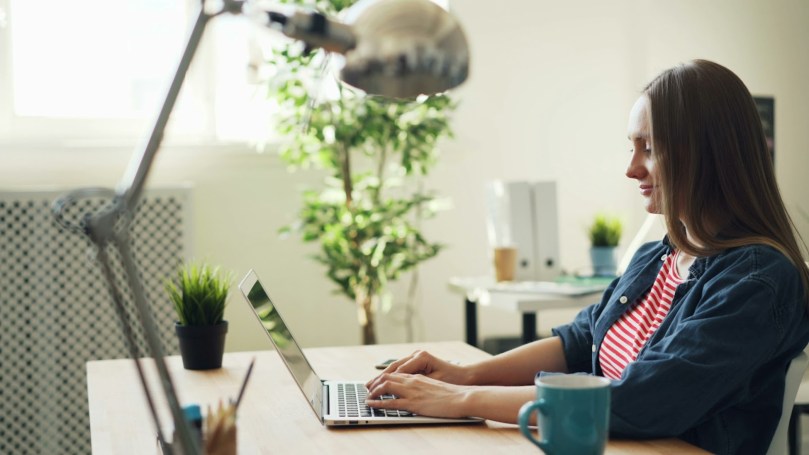 Woman types on a laptop in a well-lit room, with a small smile on her face.